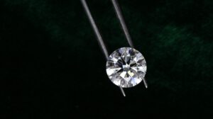 Ästhetik A sparkling round diamond is delicately held by tweezers against a dark textured background.