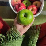 Implantologie A person holding a green apple with a bowl of mixed apples, showcasing a healthy lifestyle.